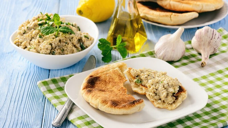 Pita-Brot mit einem Dip aus Auberginen als Brotaufstrich, daneben Knoblauch, Olivenöl und eine Zitrone