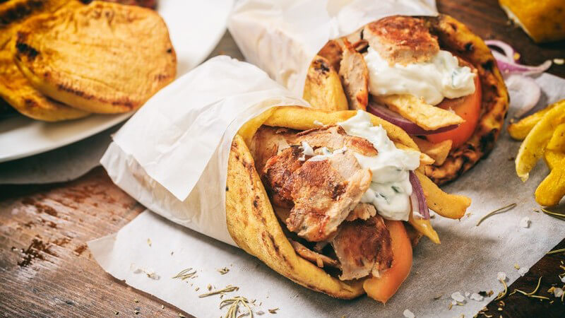 Zwei Gyros Pita Taschen in Papier eingewickelt auf Holztisch, daneben ein paar Pommes und Pita-Brote