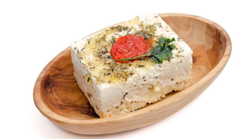 Großes Stück Feta-Käse mit Öl, Kräutern und Kirschtomate in einer ovalen Holzschale vor weißem Hintergrund