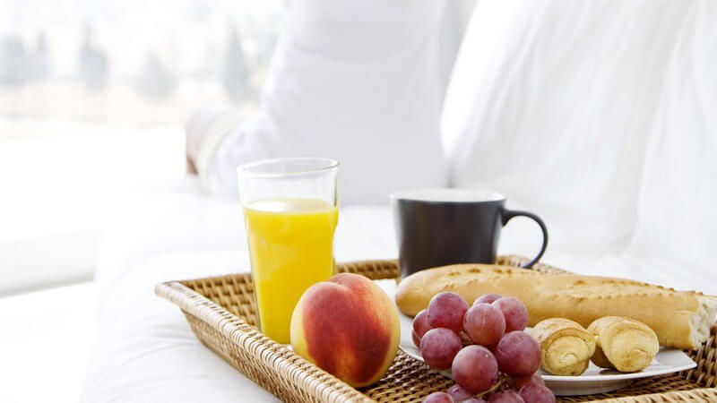 Korbtablett mit Frühstück auf weißem Bett, im Hintergrund Frau die sich streckt
