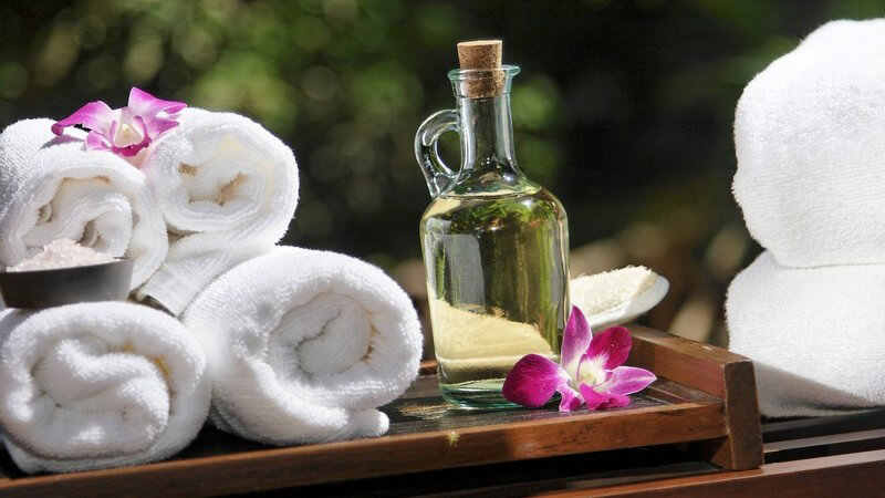Glasflasche mit Massageöl, daneben vier zusammengerollte Handtücher auf Holztablett