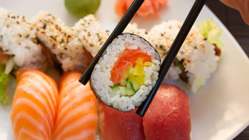 Schwarze Essstäbchen mit Sushi-Rolle über einem Teller mit weiteren Sushi-Häppchen