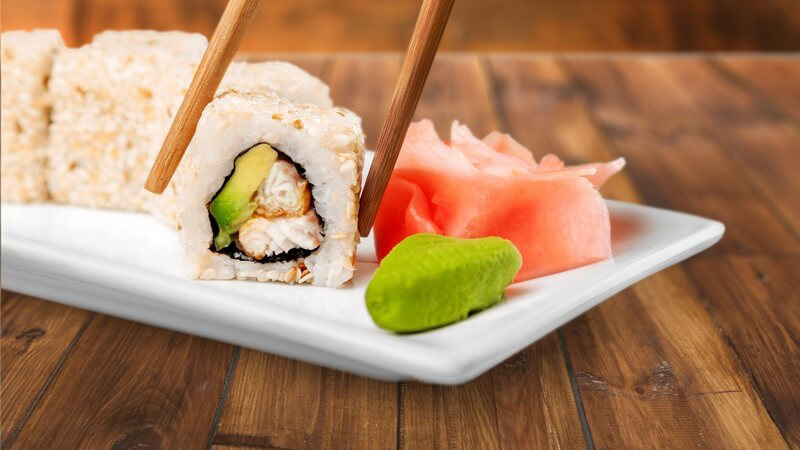 Essstäbchen greifen ein Stück Sushi von einer schmalen weißen Sushi-Platte auf Holztisch
