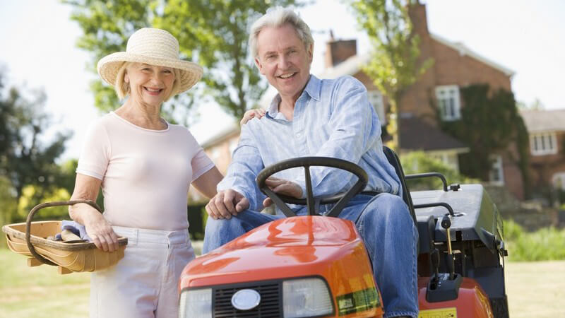 Älteres Paar im Garten, er sitzt auf Rasenmäher, sie steht neben ihm