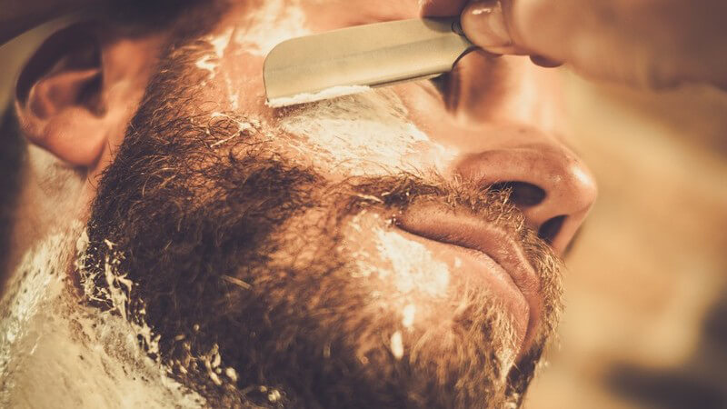 Junger Mann mit Vollbart bekommt von einem Barbier den Bart mit einem Rasiermesser abrasiert, Ansicht von unten