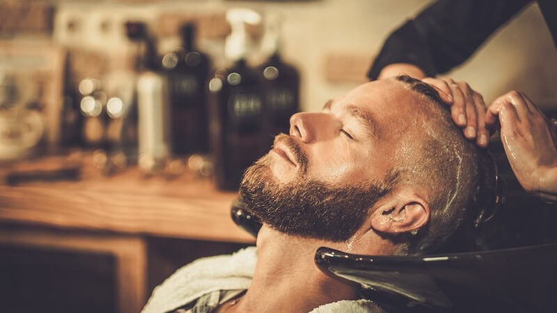 Junger Mann mit Vollbart sitzt im Friseursalon und bekommt die Haare gewaschen