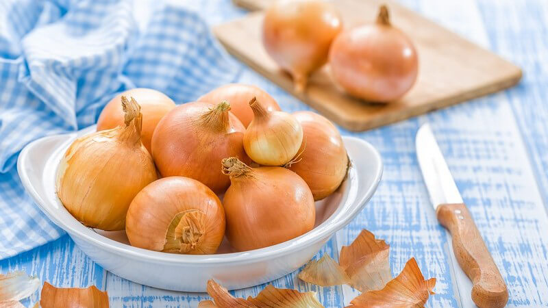 Schale mit ungeschälten Zwiebeln neben Messer und Brettchen auf blauem Holztisch