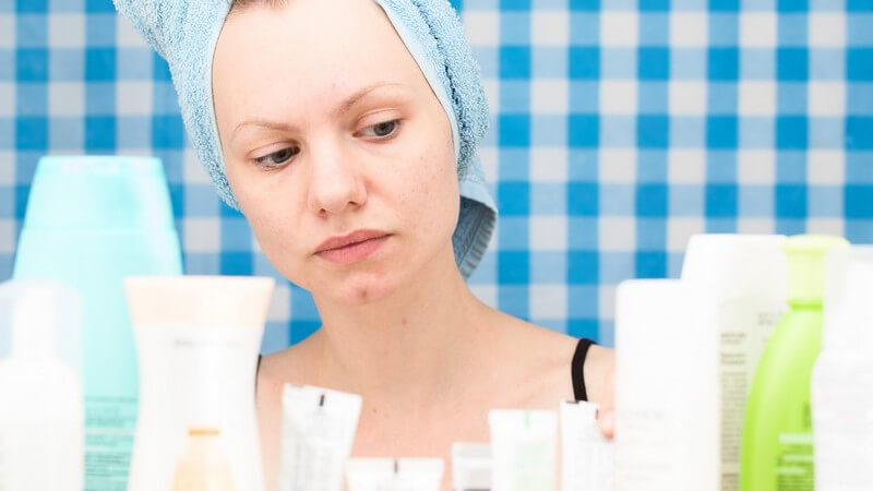 Frau mit hellblauem Handtuch im Haar guckt auf eine Ansammlung von Kosmetikprodukten, blau-weiß karierter Hintergrund