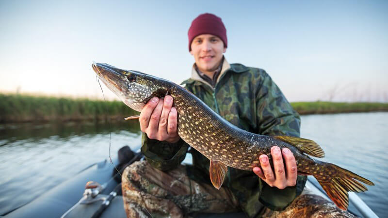 Junger Mann mit Mütze und Tarnkleidung sitzt im Fischerboot und präsentiert einen gefangenen Hecht