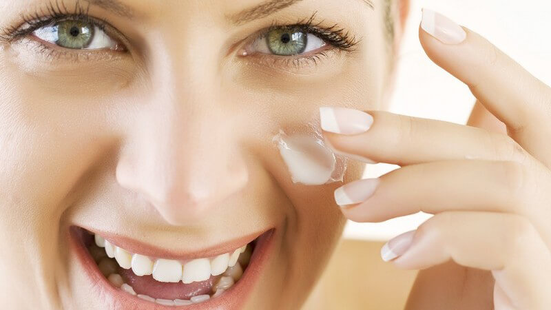 Lachende junge Frau cremt ihr Gesicht mit Creme ein