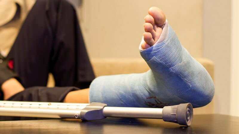 Hochgelegtes blaues Gipsbein, daneben eine Krücke auf dem Tisch