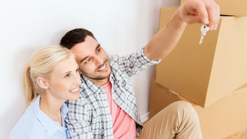 Paar sitzt an einer Wand neben Umzugskartons und hält einen Schlüssel hoch