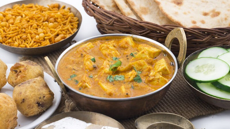 Eiserne Schüssel mit Panir (indischer Frischkäse) in brauner Soße, daneben Gurkenscheiben, Brot und andere Zutaten