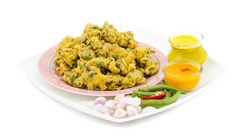 Rosa Teller mit frittiertem Gemüse (Pakora) neben Knoblauchzehen, grünen Peperoni und zwei Dips auf weißem Hintergrun