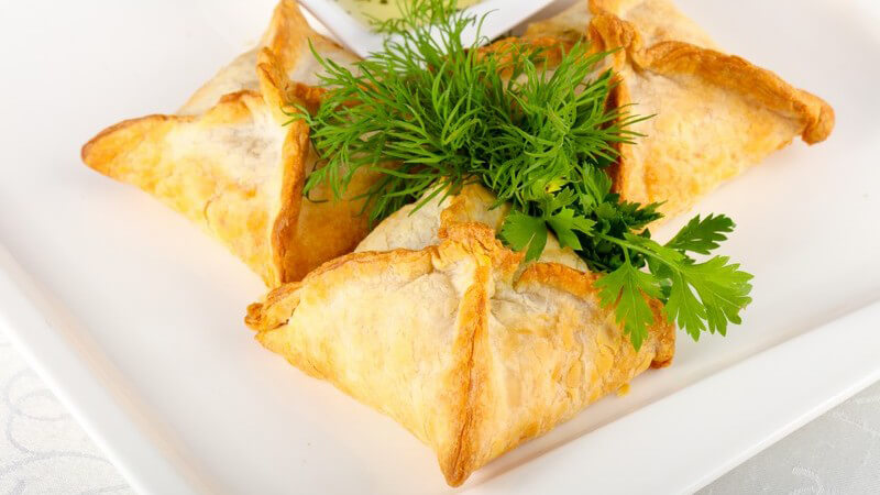 Drei viereckige Samosas (gefüllte Teigtaschen) auf weißem Teller, garniert mit grünen Kräutern