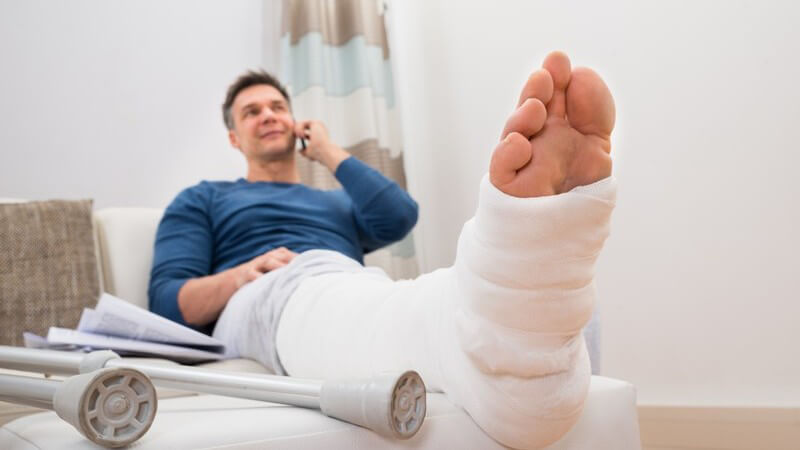 Mann mit Gipsbein liegt neben Krücken auf der Couch und telefoniert