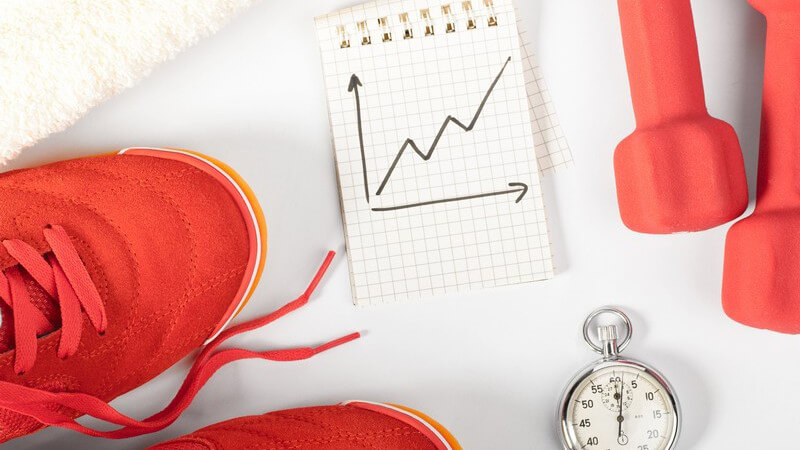Turnschuhe, Stoppuhr, Hanteln und eine Zeichnung über Trainingsfortschritte