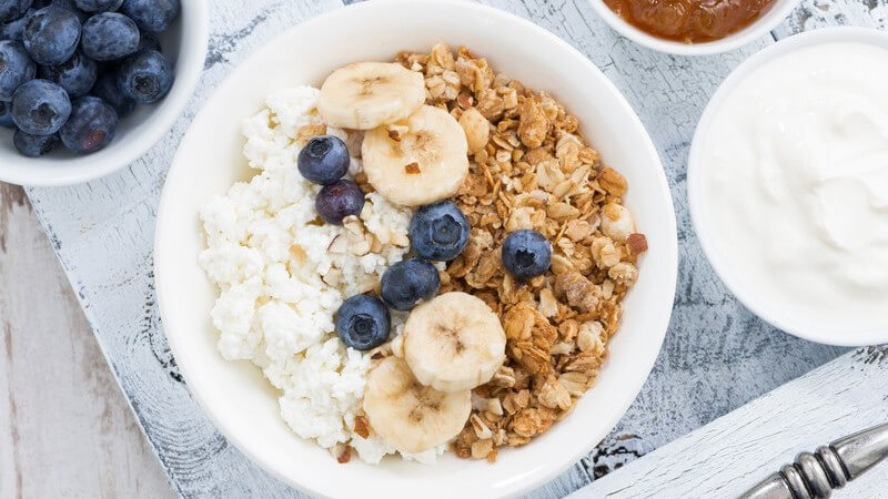 Schale mit Müsli, Hüttenkäse, Bananenscheiben und Blaubeeren, daneben Schälchen mit Blaubeeren, Joghurt und Marmelad