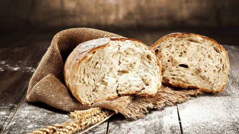 Geteilter Brotleib auf einem Jutesack, daneben etwas Getreide und Mehl