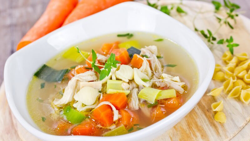 Hühnersuppe mit Möhren und Hühnchenfleisch, neben dem Teller liegen rohe Möhren und Nudeln