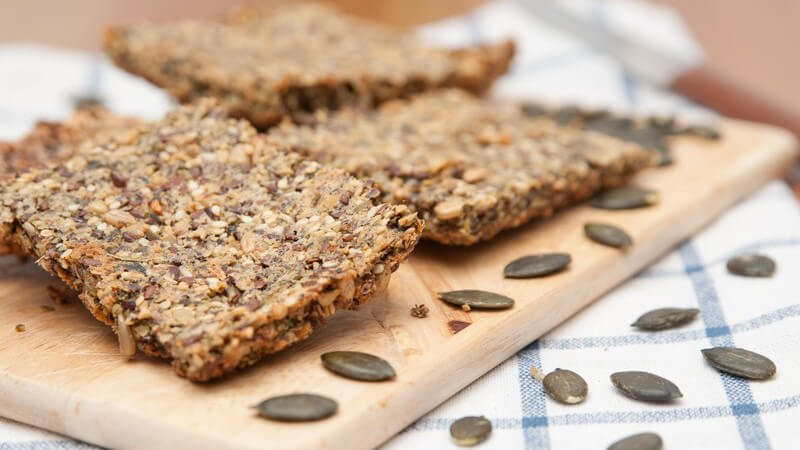 Vollkorn-Knäckebrot-Scheiben auf Holzbrett, daneben mehrere Sonnenblumenkerne