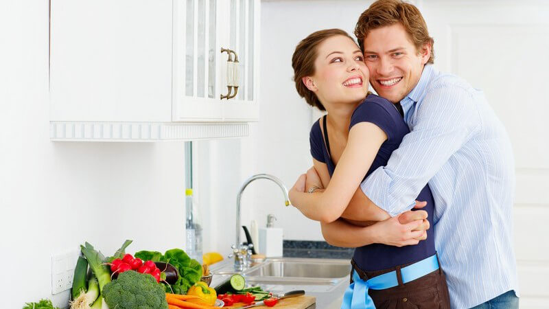 Junges Liebespaar in Küche, Umarmung, auf der Arbeitsfläche viel Gemüse, weiße Oberschränke, helle Küche, Spüle