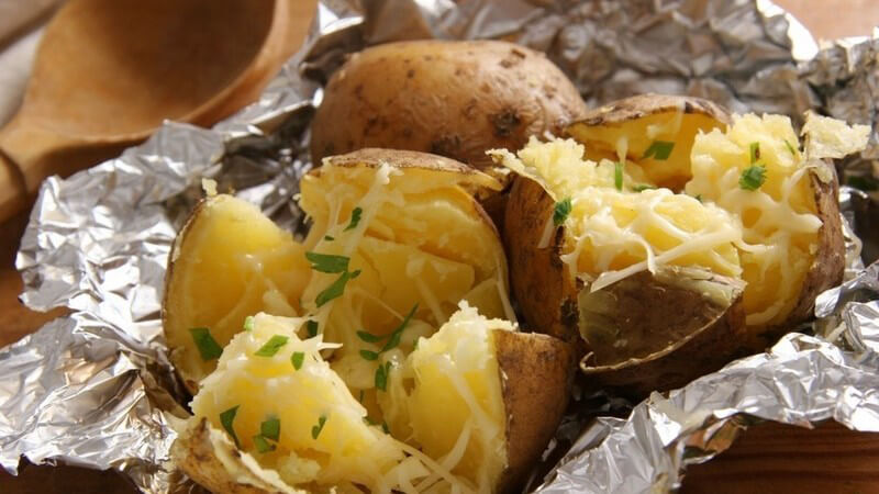 Drei offene Backkartoffeln in Alufolie mit Krautsalat und Kräutern und Holzlöffel im Hintergrund
