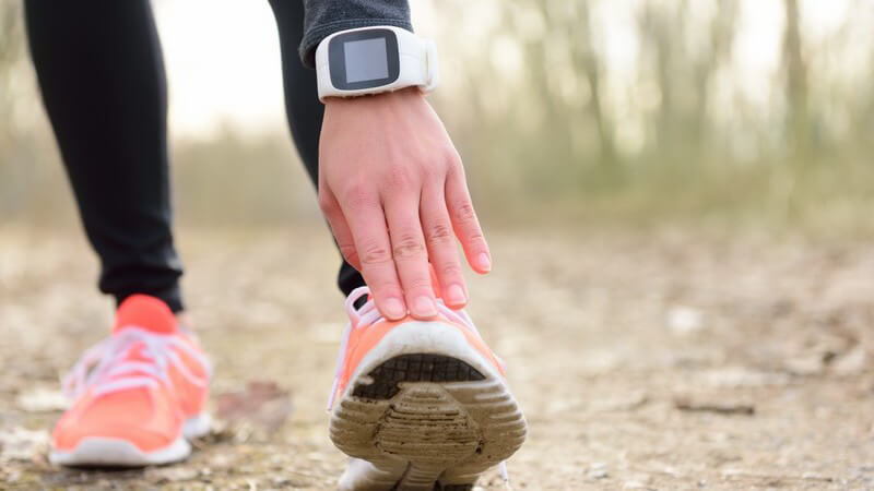 Läuferin mit weißer Smartwatch dehnt sich das linke Bein auf einem Waldweg