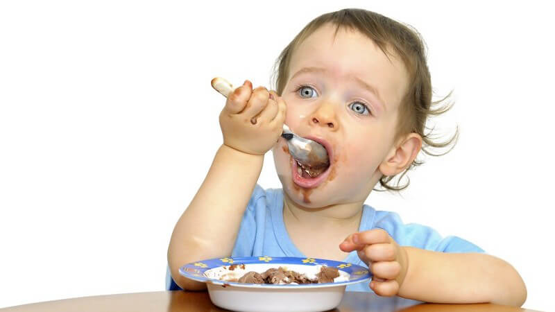 Kleinkind sitzt am Tisch und isst, Gesicht vollgeschmiert