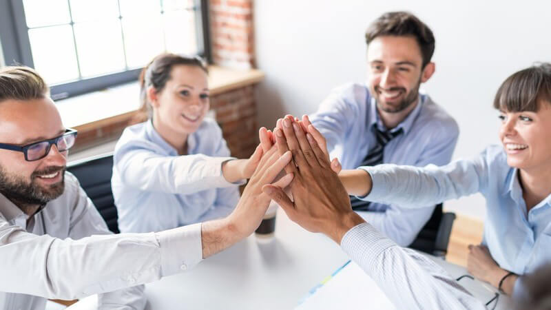 Fünf junge Geschäftsleute in blauen Hemden und Blusen sitzen am Tisch und geben sich ein High five
