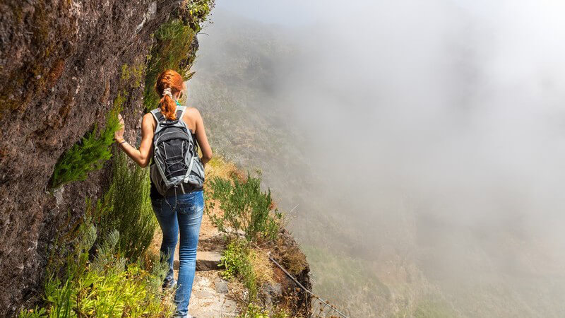 Rothaarige junge Frau mit Rucksack wandert entlang eines schmalen Pfades am Hang im nebligen Gebirge
