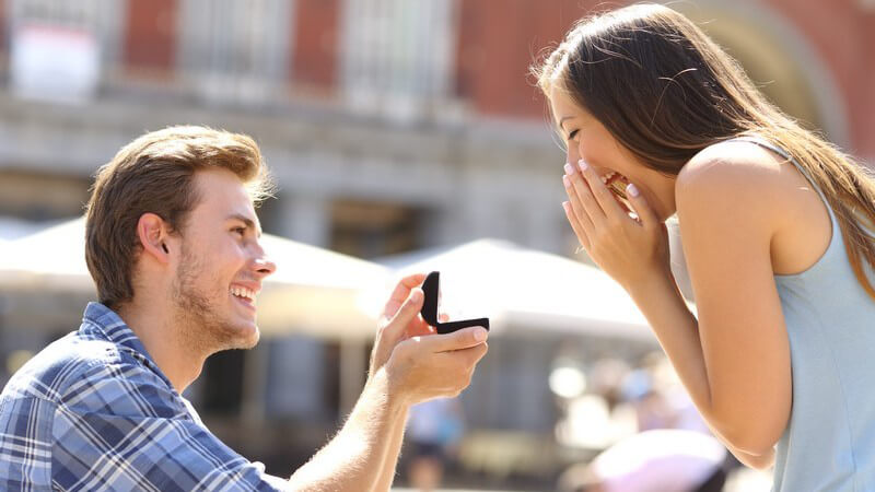 Junges Paar in einer Fußgängerzone, er kniet vor ihr nieder und hält ihr einen Verlobungsring hin
