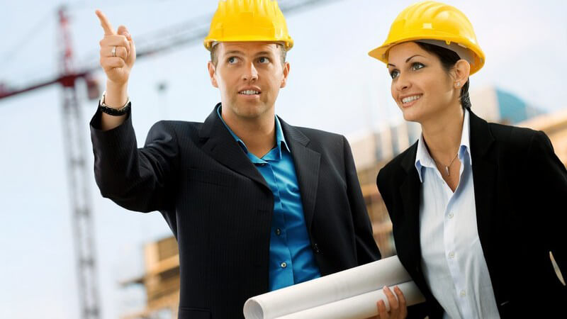 Junger Mann und junge Frau, beide in Anzug mit Bauhelm, Architekten, halten Baupläne und zeigen auf Baustelle