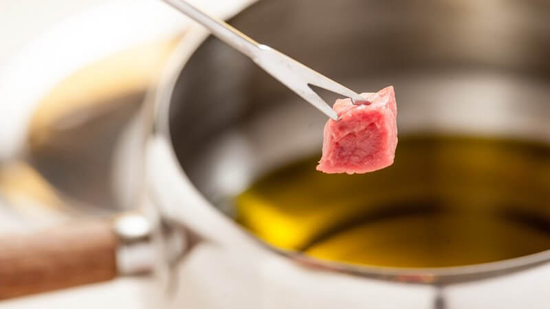 Stück Fleisch auf Fondue-Gabel, im Hintergrund Fondue-Topf mit Öl