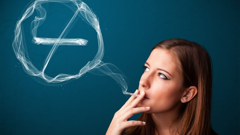 An einer Zigarette ziehende junge Frau blickt zu einer Dunstwolke in Form eines Rauchverbot-Zeichens