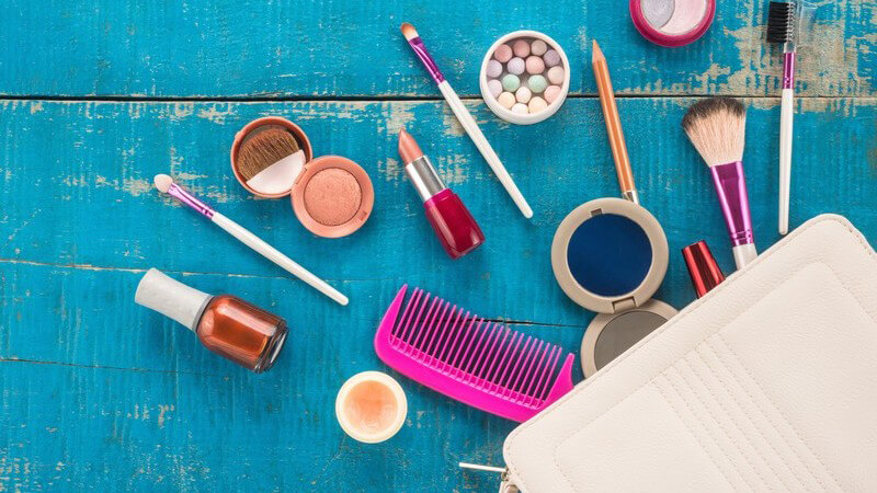 Inhalt einer hellen Handtasche wie diverses Make-up, Kamm und Spiegel liegt auf blauem Holzuntergrund