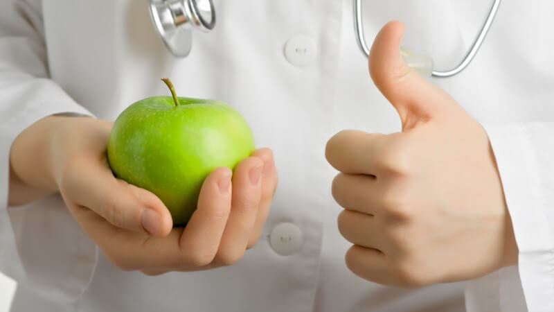 Arzt mit weißem Kittel hält grünen Apfel in einer Hand, Daumen der anderen zeigt nach oben