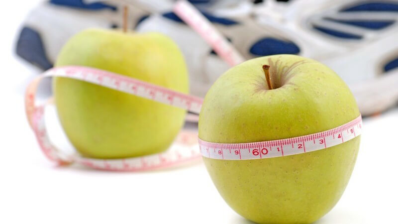 Zwei grüne Äpfel mit umgelegtem Maßband, im Hintergrund Sportschuhe