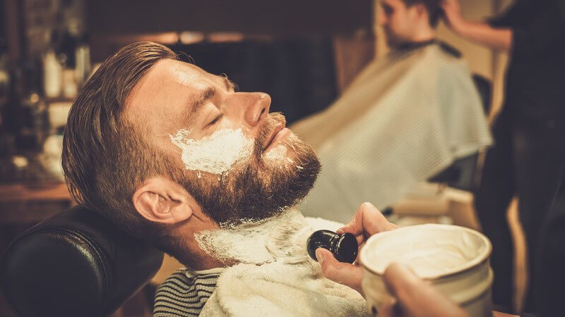 Barbier pinselt einem jungen Kunden mit Vollbart den Hals mit Rasierschaum ein