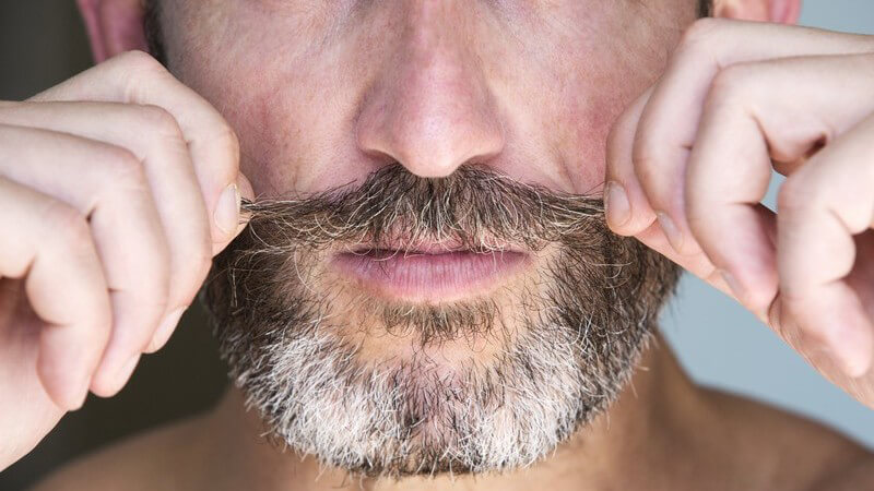 Reifer Mann mit Vollbart zieht mit beiden Händen an seinem Schnurrbart