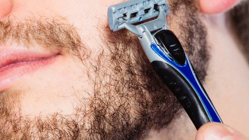 Junger Mann mit lichtem Vollbart hält einen grau-blauen Nassrasierer an seine Wange