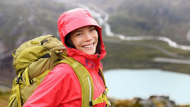 Junge Wandererin mit roter Regenjacke und Rucksack posiert im Regen vor einem Tal und Bergsee