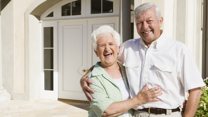 Senioren Paar steht lächelnd Arm in Arm vor Haus
