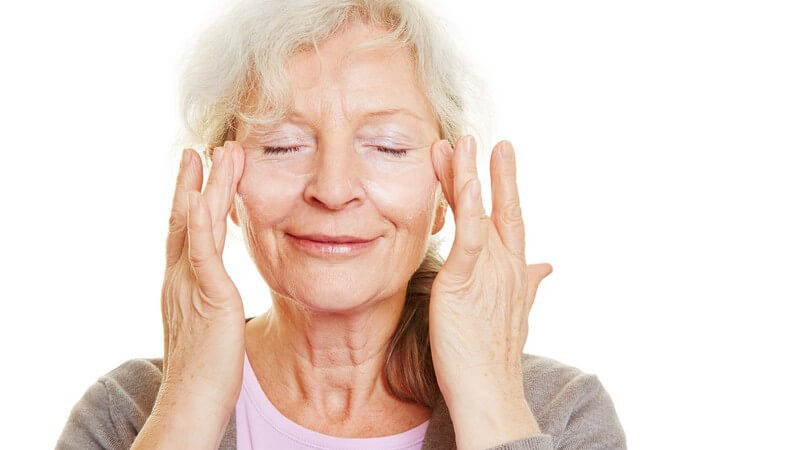 Seniorin mit langen grauen Haaren hat die Augen geschlossen und cremt ihr Gesicht ein