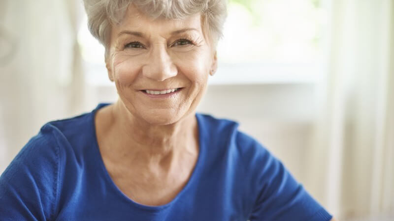 Portrait einer Seniorin mit grauen Haaren und blauem Oberteil, lächelt in die Kamera