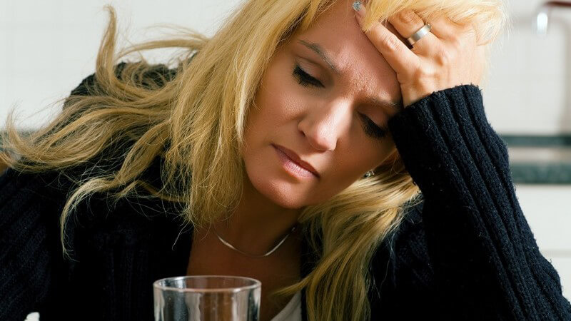 Blondierte, braune, stark geschminkte Frau, hält sich Kopf, vor ihr ein Wasserglas, Kopfschmerzen, Kater, Depression