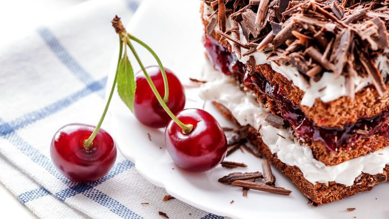 Stück Schwarzwälder Kirschtorte neben drei Kirschen am Stängel