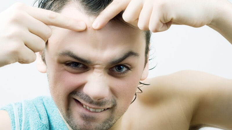 Junger Mann vor Spiegel drückt Pickel auf Stirn aus