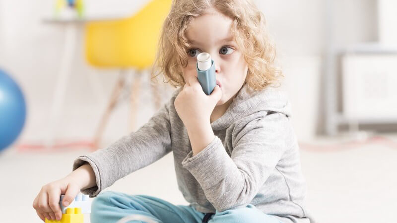 Mädchen sitzt auf dem Boden und spielt mit Bausteinen während sie aus einem Inhalator inhaliert