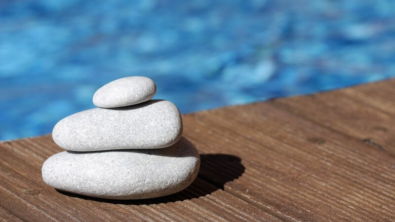 Drei aufeinander leigende Steine auf Holzuntergrund, im Hintergrund Wasser